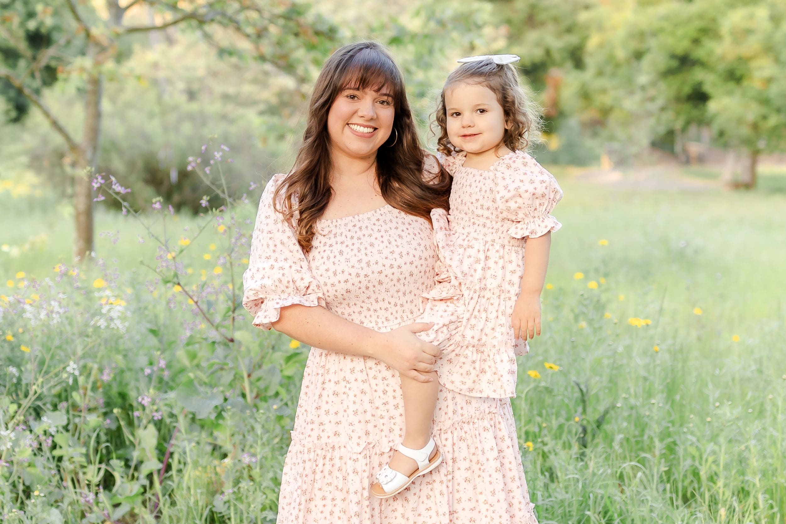 A mother and daughter in matching pink dresses pose together in a wildflower meadow during a spring photo session.