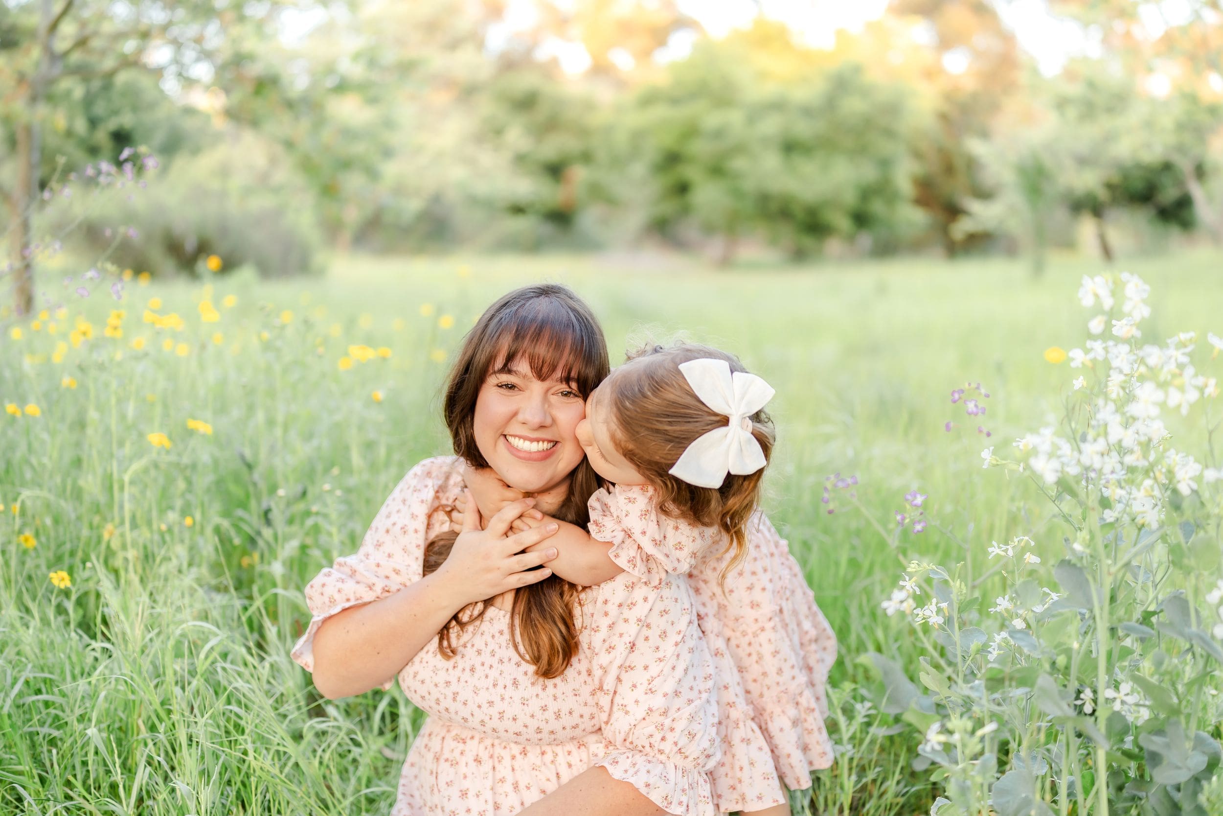 Two people in matching pink floral dresses sit together in a sunny meadow with wildflowers and greenery in Orange County, Ca