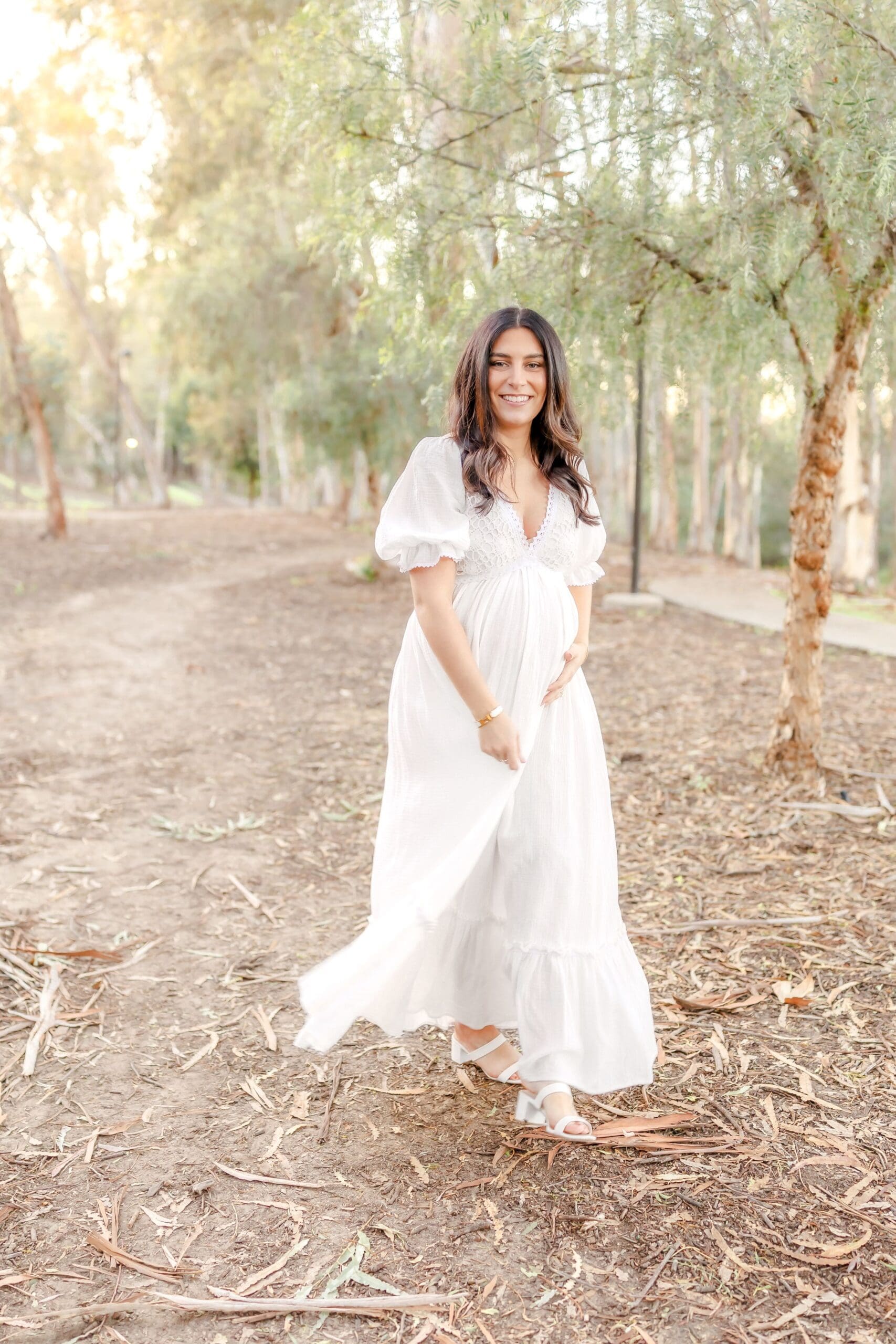 A person in a flowing white dress poses gracefully in a sun-dappled eucalyptus grove during a photoshoot.