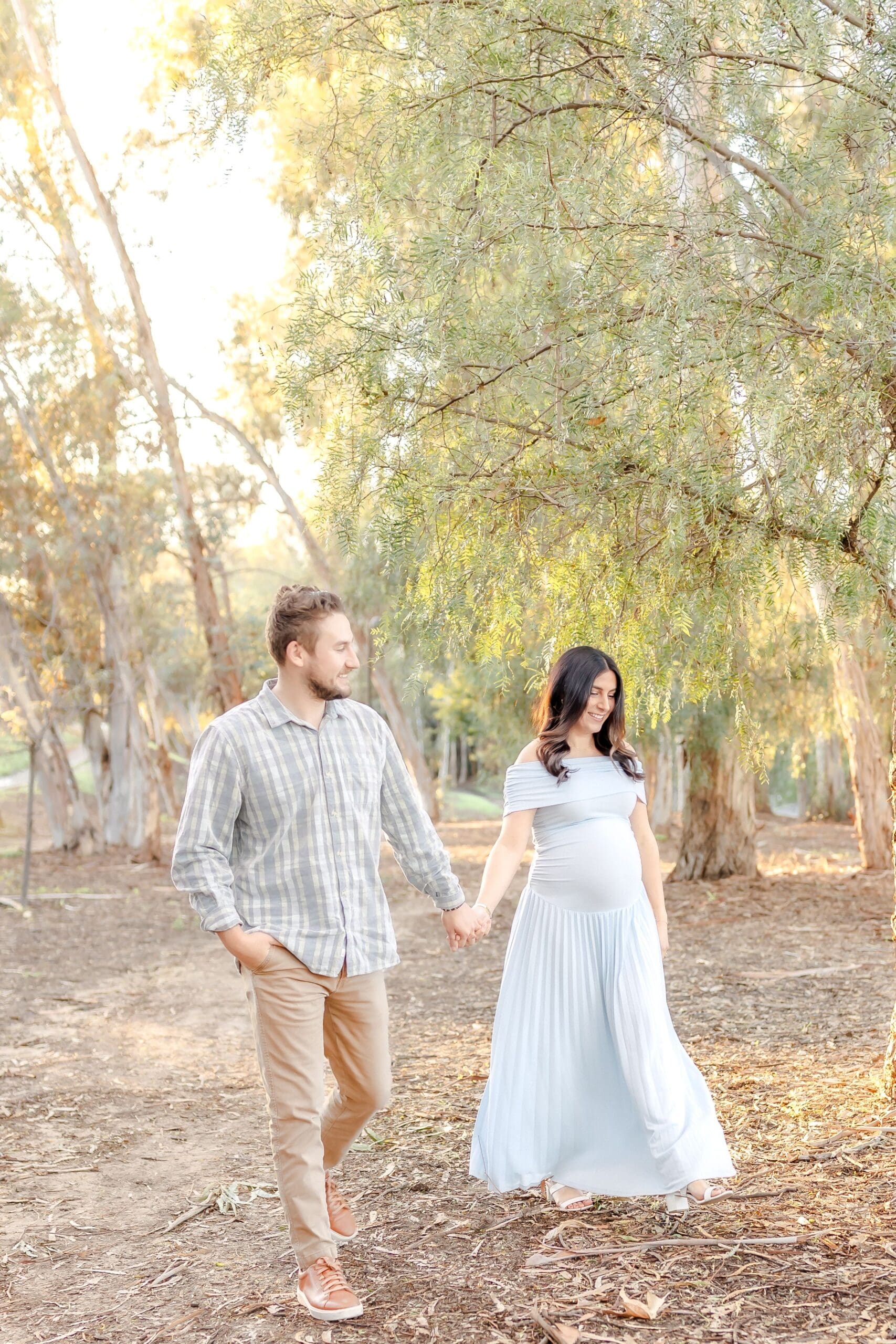 A couple in white outfits shares romantic moments while walking through a sun-dappled grove of willow trees.