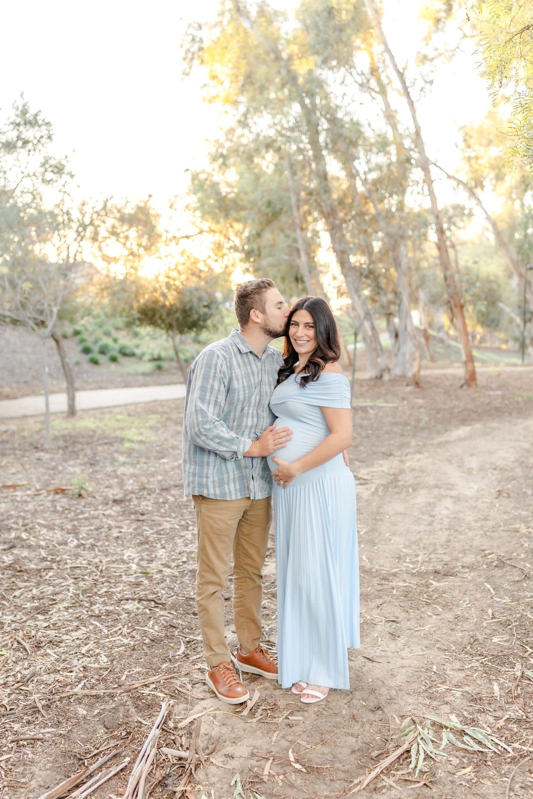 A couple shares tender moments in a sunlit wooded area wearing light blue and khaki casual outfits.