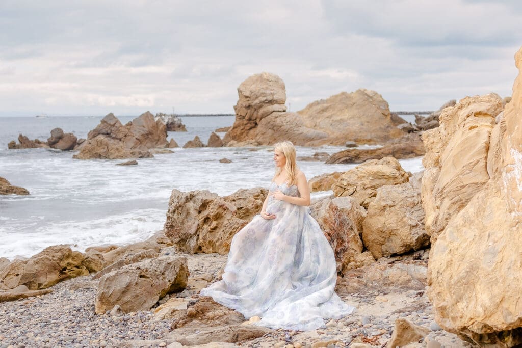 Expecting mother sitting on the rocks looking out onto the ocean during a luxury maternity session