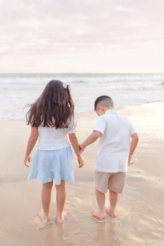 Siblings holding hands while walking along the shoreline at sunset in Newport Beach.